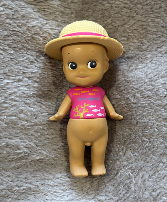 A Cute Sonny Angel Caribbean Series Tan mini figure kewpie doll with Pink Top and Straw Hat lying on grey blanket 