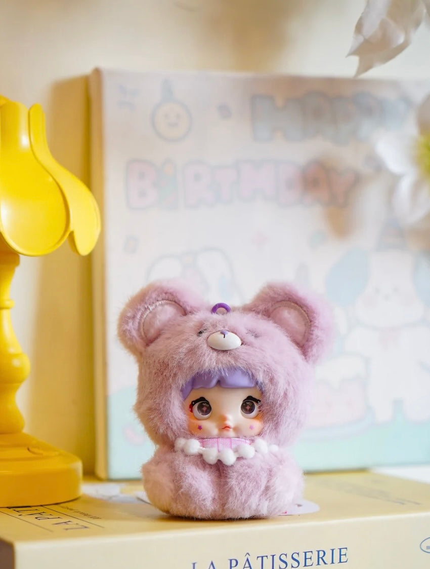 Small plush nommi baby V1 with a bear costume on a surface with a blurred background